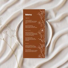 a menu on a plate next to some flowers and a white cloth with a brown border