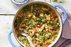 a blue pot filled with pasta and spinach on top of a wooden table next to a spoon