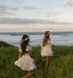 two girls in white dresses walking through tall grass near the ocean