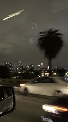 cars driving down the road at night with city lights in the background and palm trees