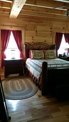 a bed sitting in the middle of a bedroom next to two windows with red curtains