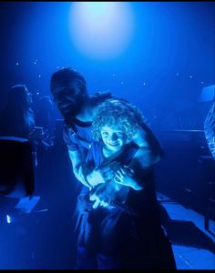 two people are hugging in the dark on stage with blue lights behind them and one person is holding his arm around the other man's shoulder