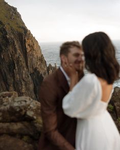 a man and woman standing next to each other on top of a cliff near the ocean