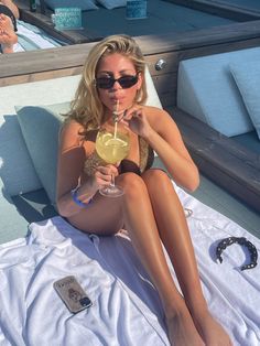 a woman is sitting on the beach with a drink in her hand and wearing sunglasses