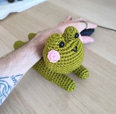 a hand holding a green crocheted frog on top of a wooden table next to a computer mouse