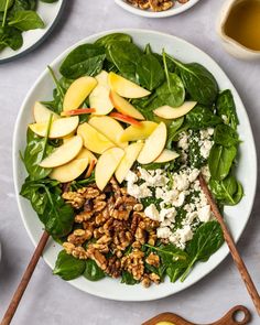 a white plate topped with spinach, apples and walnuts next to bowls of nuts