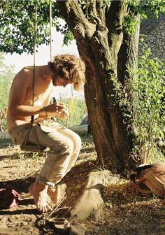 a shirtless man sitting on a swing next to a tree and looking at his cell phone