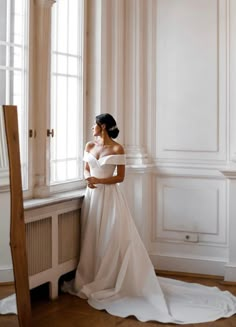 a woman standing in front of a window wearing a wedding dress and looking out the window