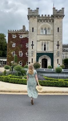 a woman is walking down the street in front of a castle like building with hedges