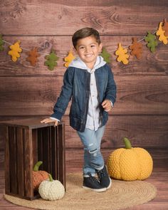 a young boy standing next to a wooden box and pumpkins on a rug in front of a wood wall