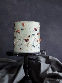 a white cake with flowers on it sitting on top of a black stand next to a gray wall