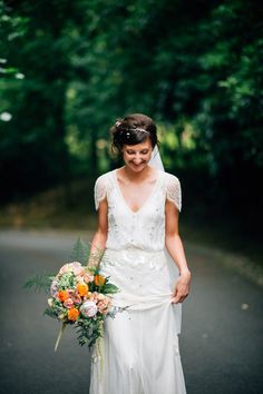 a woman in a white dress holding a bouquet and walking down the road with trees behind her