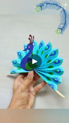 a hand holding a paper peacock on top of a white table next to a blue feather