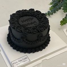 a black frosted birthday cake sitting on top of a white box next to a plant