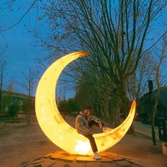 a man sitting on top of a large yellow crescent shaped object in the middle of a park