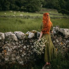 a woman with long red hair is standing by a stone wall holding a bouquet of flowers