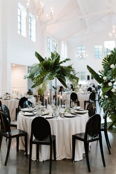 a table set up with white linens, black chairs and greenery in the center