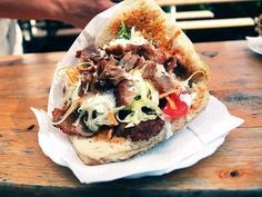 a sandwich with meat and vegetables on it sitting on top of a wooden table next to a person