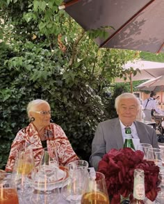 two people sitting at an outdoor table with wine glasses and flowers in front of them