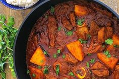 a pot filled with stew and vegetables on top of a wooden table next to rice