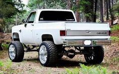 a white truck parked in the woods with large tires on it's front wheels