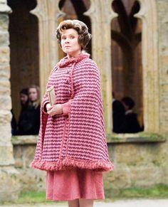 a woman in a pink coat and dress is standing near an old building with people looking on