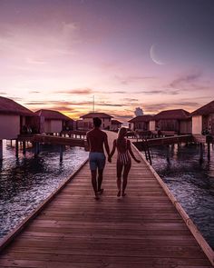 two people walking down a pier towards the water at sunset or dawn with houses in the background