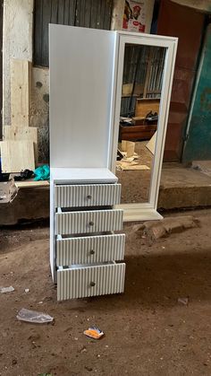 a white dresser sitting on top of a dirt floor next to a mirror and trash