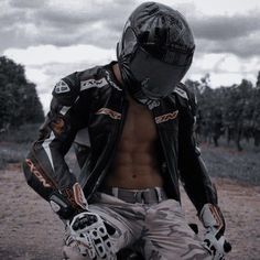 a shirtless man in black jacket and white pants sitting on a dirt road with his motorcycle gear