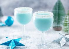 two glasses filled with blue liquid sitting on top of snow covered ground next to christmas trees