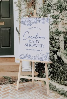 a baby shower sign sitting in front of a door with greenery on the side