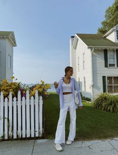 a woman standing in front of a white picket fence with flowers on the grass and houses behind her