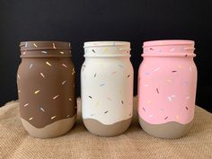 three painted jars with sprinkles are sitting on a burlap surface