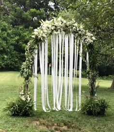 an outdoor wedding ceremony with white flowers and ribbons hanging from the arch, surrounded by lush greenery