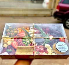 a person holding up a box filled with fruit and crackers in front of a car