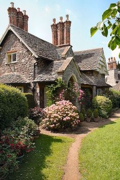 a stone house with lots of flowers in the front yard and bushes on either side