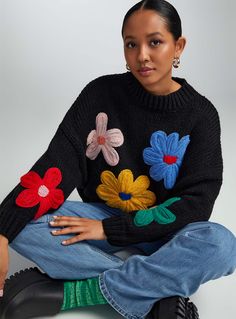 a woman sitting on the ground wearing a black sweater with colorful flower appliques