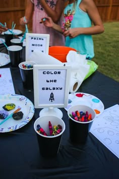 two girls are standing behind a table with cups and plates on it that have crayons in them