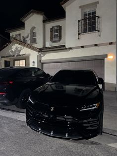 two black sports cars parked in front of a white house at night with lights on