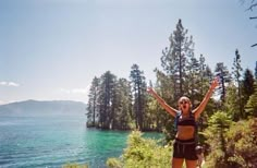 a woman standing on the edge of a cliff with her arms in the air and trees around her