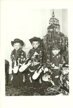 black and white photograph of three boys dressed up in cowboy outfits with christmas tree behind them