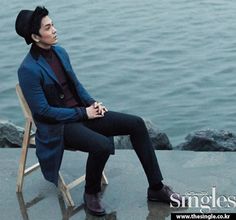 a man is sitting on a chair by the water wearing a blue coat and black hat