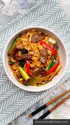 a white bowl filled with noodles and meat on top of a blue table cloth next to chopsticks