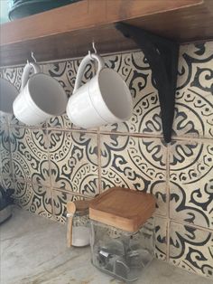 three coffee mugs hanging on the wall above a wooden stool in a tiled kitchen