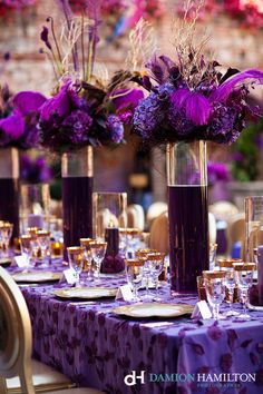 purple flowers are in tall vases with gold rimmed glasses and place settings on the table