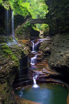 a small waterfall in the middle of a forest with a bridge over it and water running underneath