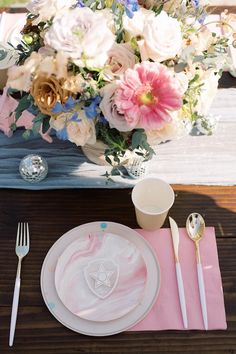 a table setting with pink and white plates, silverware and flowers in the background