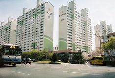 two buses are driving down the street in front of tall buildings with green and white designs