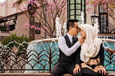 a man and woman sitting on a bench in front of a fountain with pink flowers