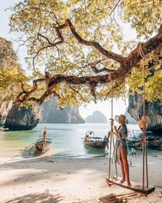 a woman is standing on a swing by the beach with boats in the water behind her